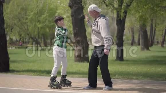 可爱的男孩穿着格子衬衫在公园里滑旱冰他的祖父抓住他拥抱主动休闲视频的预览图