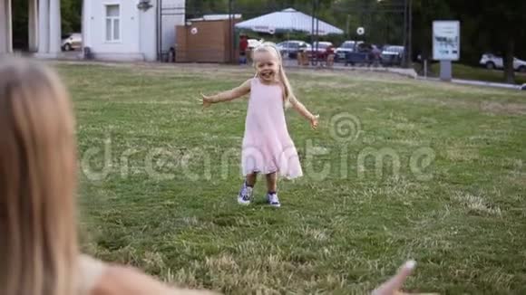 一个漂亮的金发女孩穿着粉红色的连衣裙在草坪上追赶她的母亲一位成年母亲在公园里和她玩耍视频的预览图