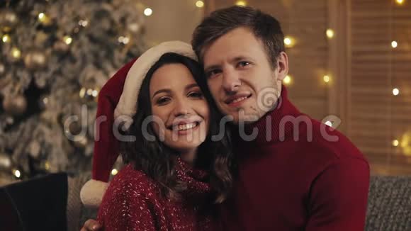 一对幸福微笑的夫妇坐在靠近圣诞树的客厅的沙发上看着镜头的肖像概念视频的预览图