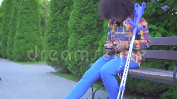 非洲裔美国妇女发型为非洲发型腿骨折拄着拐杖坐在长凳上身体紧闭视频的预览图