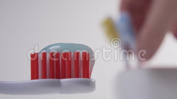 白色牙刷上面有红色刚毛和蓝色牙膏在背景中一只手从玻璃杯中拿起一支蓝色牙刷视频的预览图