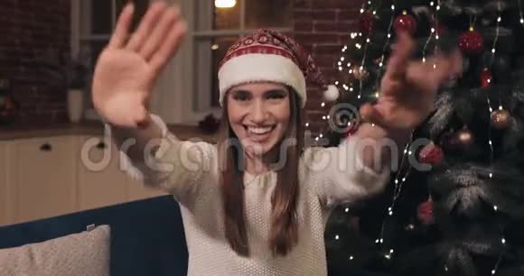 戴着圣诞帽的快乐白种人漂亮女人坐在家中的圣诞树旁挥舞着喜献红礼物盒视频的预览图