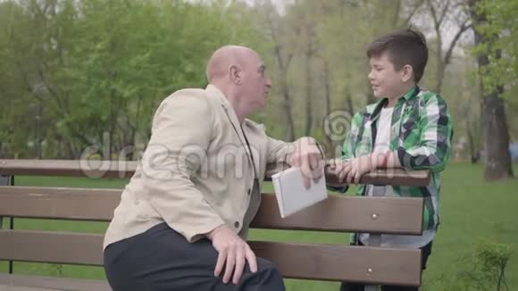 那个秃顶的成熟男人拿着一本书坐在公园的长凳上和他的孙子说话然后人们握手视频的预览图