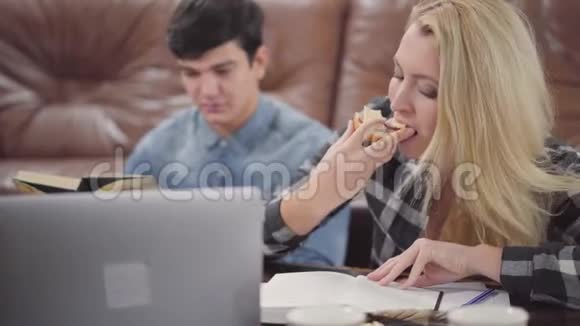 肖像可爱的金发年轻女子在看笔记本时吃三明治而她的男朋友在探索有关信息视频的预览图