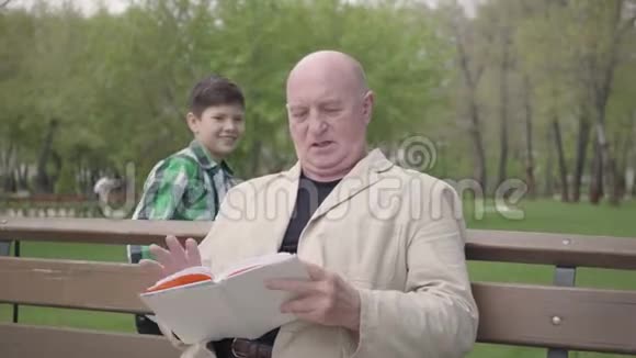 肖像可爱的秃头成熟男人坐在公园的长凳上看书可爱的小男孩从后面走来视频的预览图