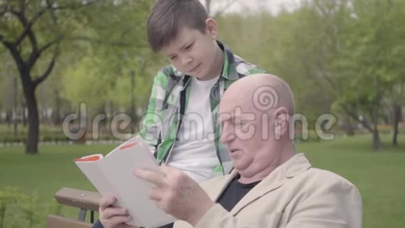 肖像可爱的爷爷和可爱的孙子坐在公园的长凳上老人为男孩读这本书视频的预览图