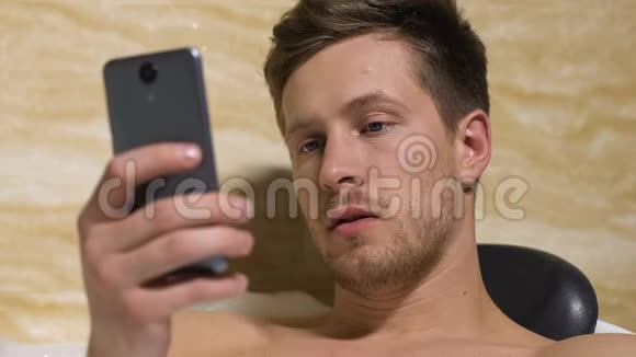 无聊的年轻人在浴缸里滚动电话阅读乏味的信息应用程序视频的预览图