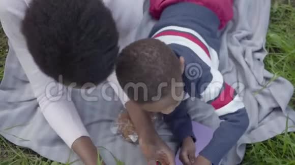 可爱的非裔美国妇女短发躺在公园的草地上儿子玩塑料变压器玩具视频的预览图