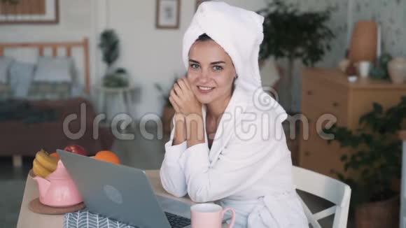 头上戴毛巾的女孩坐在厨房里手提电脑看着相机微笑慢动作视频的预览图
