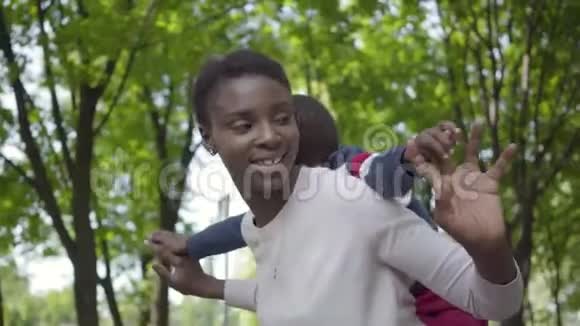 可爱的非洲裔美国妇女抱着儿子背在绿色公园里的肖像手臂向旁边靠近视频的预览图