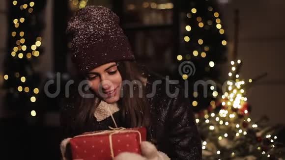 穿着冬帽和戴着红色礼物盒的米滕斯笑着打扮的女孩女人看着幸福站在灯光下视频的预览图