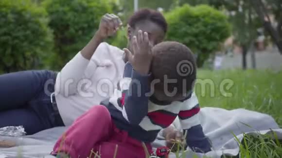 肖像可爱的非裔美国妇女躺在毯子上她的小儿子在公园里小妈妈挠痒痒男孩视频的预览图