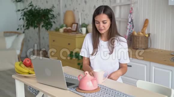 女孩在笔记本电脑上工作从水壶里倒茶手里拿着杯子动作缓慢视频的预览图