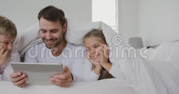 父亲和孩子们在家里的床上用毯子盖着数字平板电脑视频的预览图