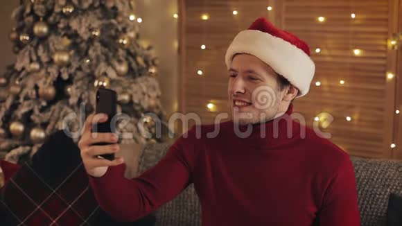穿着圣诞老人帽子在圣诞树附近的沙发上打坐的白种人用他的智能手机视频的预览图