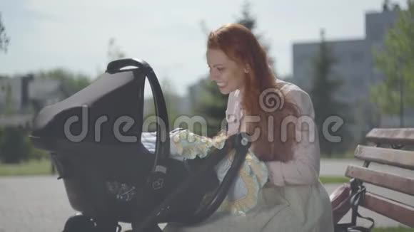 年轻快乐的红头发母亲坐在房子附近的长凳上坐着婴儿车微笑着把他拉到一个漂亮的椅子上视频的预览图