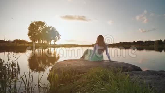 一个孤独的女孩坐在夕阳背景下的石头上的一个湖上录像在移动视频的预览图
