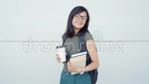 一个快乐的学生女孩在灰色背景下背着背包书和一杯咖啡的肖像视频的预览图