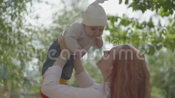 一位年轻美丽的红头发母亲在春天阳光明媚的阳光下抱着她可爱的小儿子在街上玩耍视频的预览图