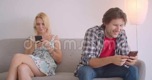 一对有魅力的白种人成年夫妇使用电话和分享坐在室内沙发上的滑稽帖子的特写照片视频的预览图