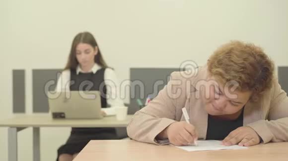 紧张的年轻丰满的女人写下信息坐在现代办公室的前景而她的苗条漂亮视频的预览图