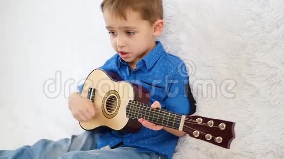 一个快乐的孩子坐在白色的沙发上弹着孩子们吉他和唱歌视频的预览图