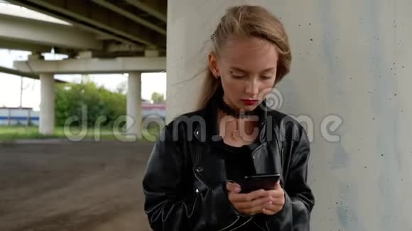 穿黑色皮夹克的年轻女孩在城市街道上使用手机在车桥下拿着智能手机的摇滚女孩视频的预览图