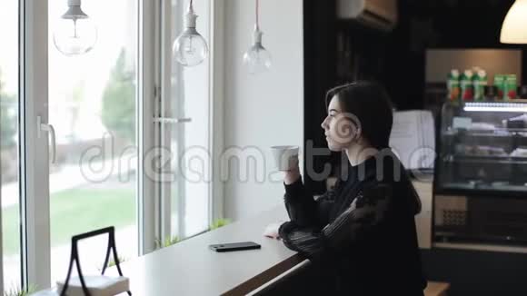一个漂亮的年轻女孩坐在咖啡店里喝着一杯茶用她的智能手机侧视视频的预览图