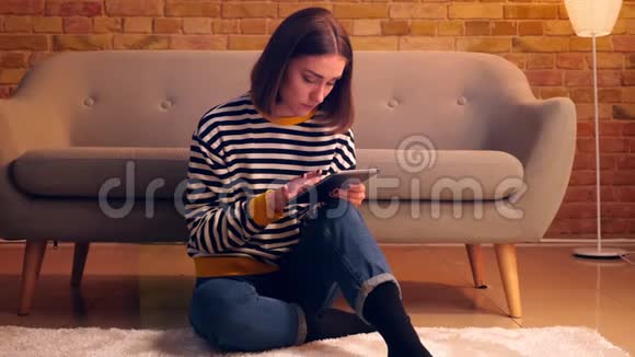在室内舒适的公寓里一个年轻漂亮女孩用平板电脑坐在地板上的特写肖像视频的预览图