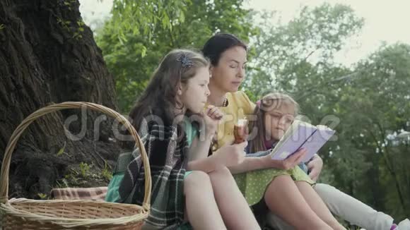 成熟的祖母坐在靠近树的公园里躺在毯子上和两个吃着美味的孙女一起看书视频的预览图