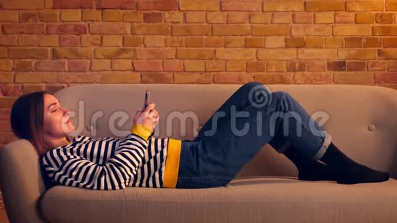 一个年轻漂亮的女孩用电话和微笑躺在舒适的室内公寓沙发上的特写照片视频的预览图