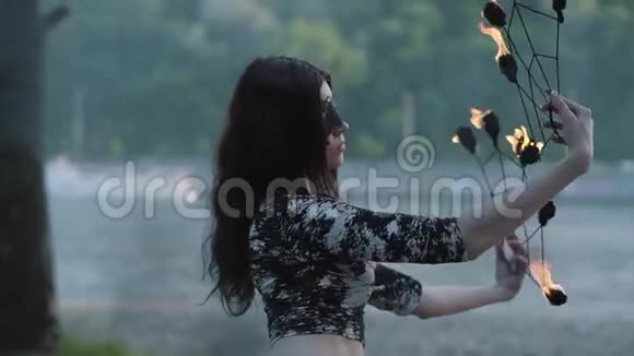 戴着面具的年轻漂亮女人在树前的河岸上表演火焰表演的肖像技能视频的预览图