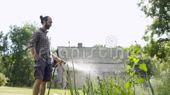 年轻英俊的嬉皮士用花园软管浇灌花园视频的预览图