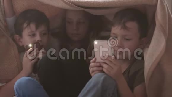 两个兄弟姐妹坐在毯子下面一起玩手机小兄弟在家里消磨时间视频的预览图