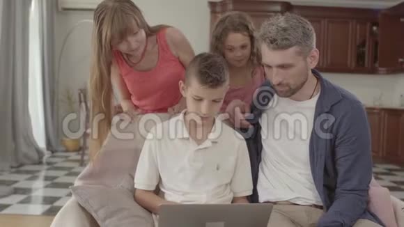 父亲和儿子一起坐在扶手椅上拿着笔记本电脑美丽的母女站在后面视频的预览图