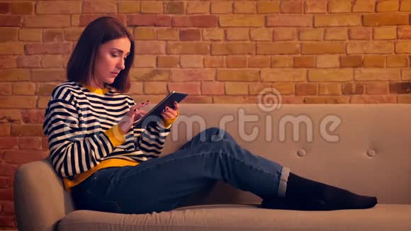 在室内舒适的公寓里一个年轻漂亮女孩用平板电脑坐在沙发上的特写画像视频的预览图