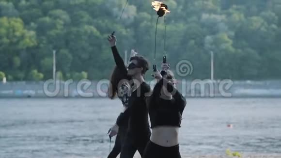 有技巧的男人和两个穿着黑色衣服和面具的女人站在河岸上用火焰表演节目技能视频的预览图