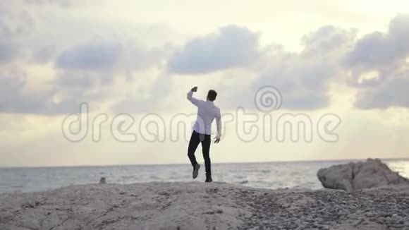 年轻的快乐男子在沙滩上向水中扔石头欣赏令人惊叹的海滨景色和美丽的天气视频的预览图