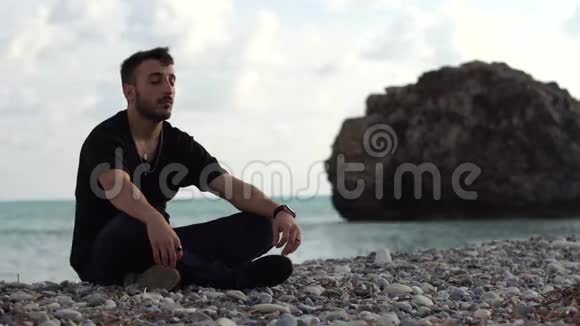 英俊的男人在岩石和大海的背景上沉思时光飞逝一个闭着眼睛坐着的年轻人视频的预览图