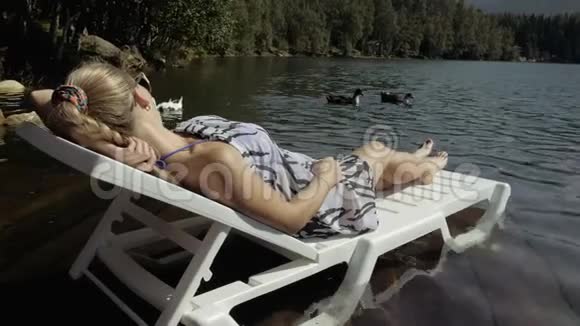 女人躺在日光浴床上戴着太阳镜和一条薄纱披肩女孩在洪水木水下码头休息鸭子和天鹅视频的预览图