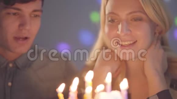 特写肖像可爱的微笑金发女孩戴着生日帽吹灭蛋糕上的蜡烛年轻人坐在附近视频的预览图