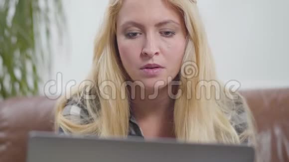 肖像可爱的金发快乐的微笑女孩坐在笔记本电脑前的皮革沙发上看着笔记本电脑屏幕视频的预览图