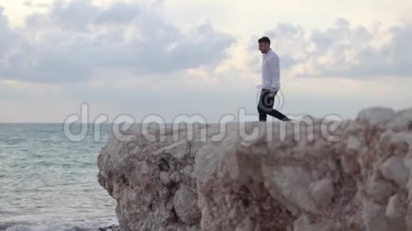 年轻的快乐男人坐在高高的岩石山上欣赏着令人惊叹的海滨景色和美丽的天气塞浦路斯帕福斯视频的预览图