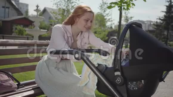 肖像美丽的正面红发女人微笑着看着坐在长凳上的公园里的婴儿车女士视频的预览图