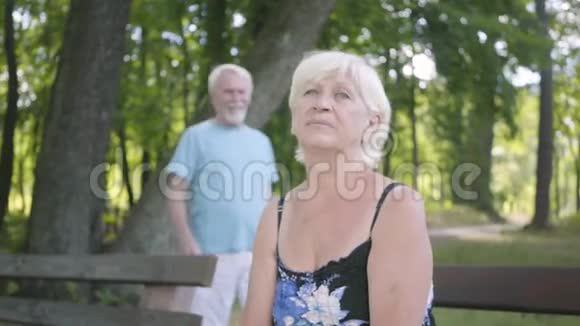 公园里坐在长凳上的可爱成熟女人的肖像微笑的高级男人从她身后走过来掩护视频的预览图