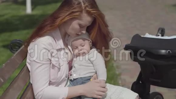 肖像可爱的红发女人和坐在长凳上的孩子玩站在附近的婴儿车女士视频的预览图