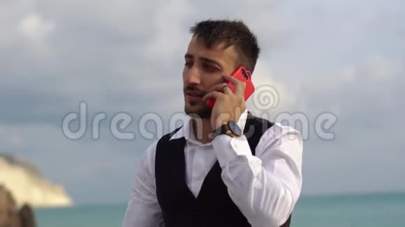 年轻可爱的时尚男人在靠近惊人的海景的牢房交谈商人在户外用手机说话视频的预览图