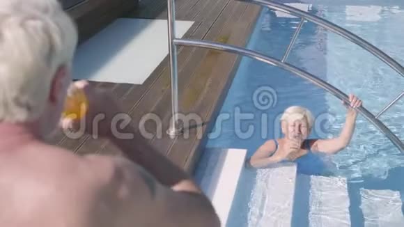 后景成熟的男人喝着果汁看着妻子在游泳池里游泳成熟的情侣在游泳池里放松视频的预览图