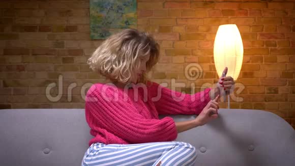 金发家庭主妇穿着粉红色毛衣拥抱她的腿在舒适的家庭氛围中在智能手机上制作美丽的自拍视频的预览图