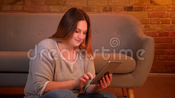 超重的长发女性自由职业者坐在地板上在舒适的家庭氛围中愉快地与平板电脑合作视频的预览图